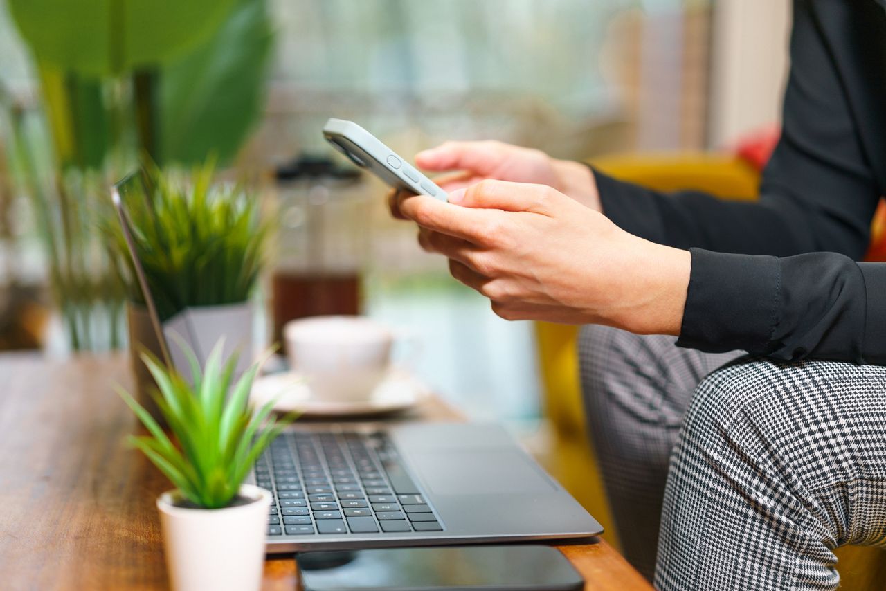 Portfolio A person using a smartphone in a cozy home office setting with plants, a laptop, and coffee.